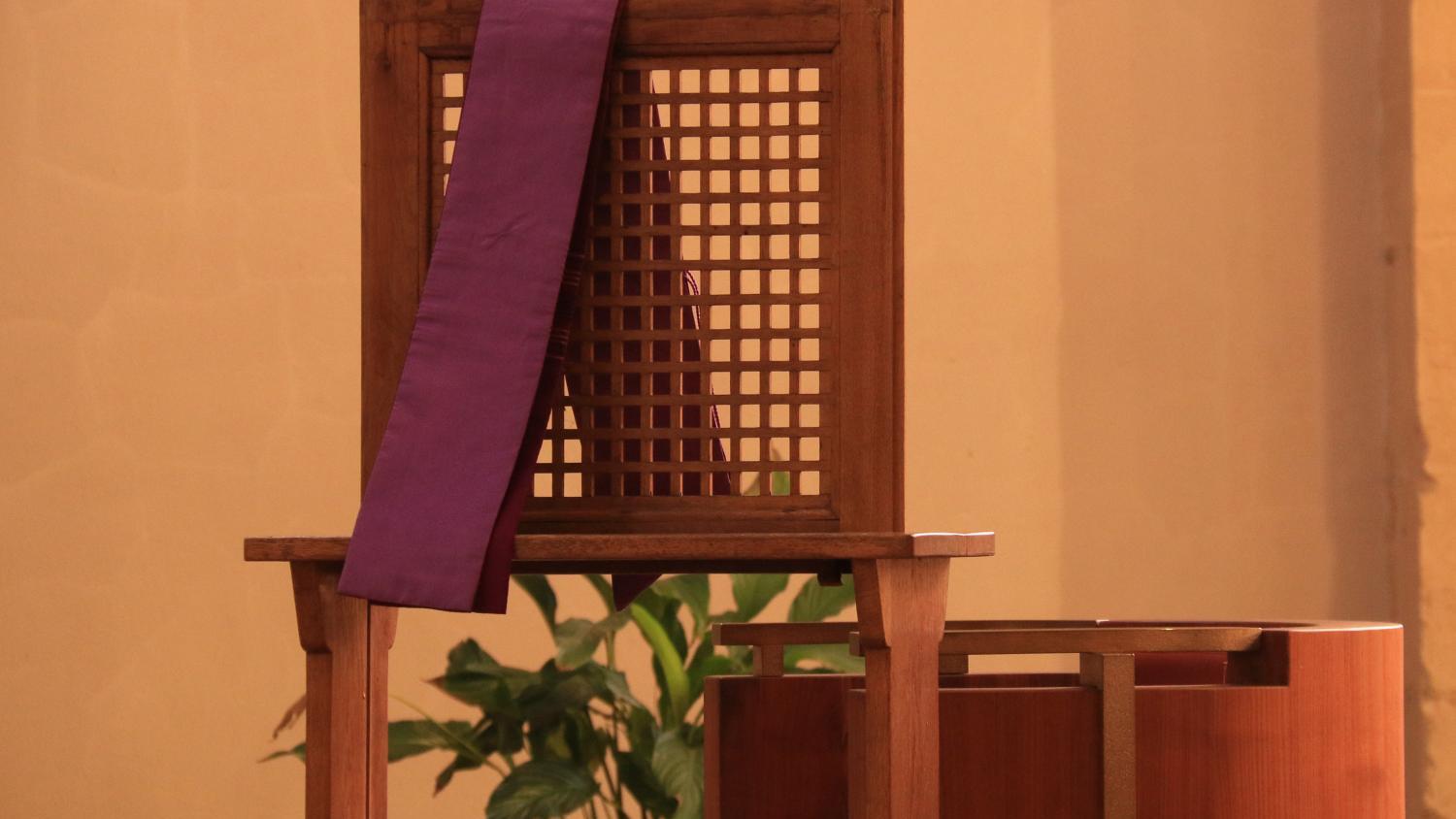 Foto eines Beichtstuhls aus Holz, über der Lehne hängt eine violette Schärpe