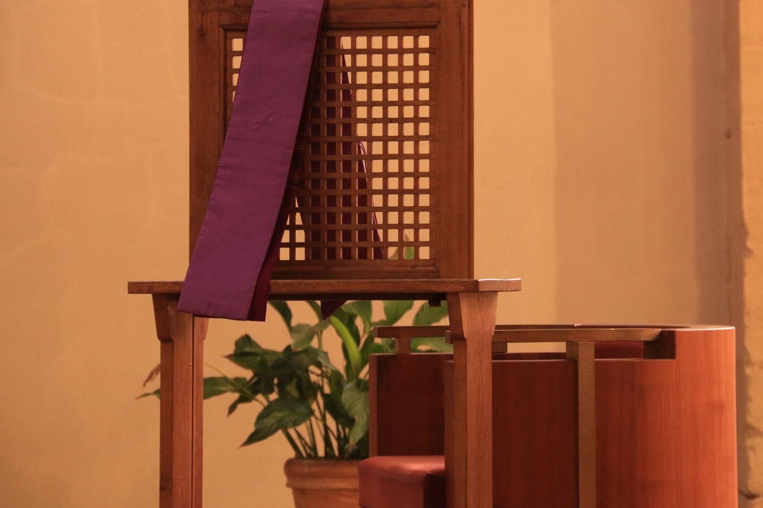 Foto eines Beichtstuhls aus Holz, über der Lehne hängt eine violette Schärpe