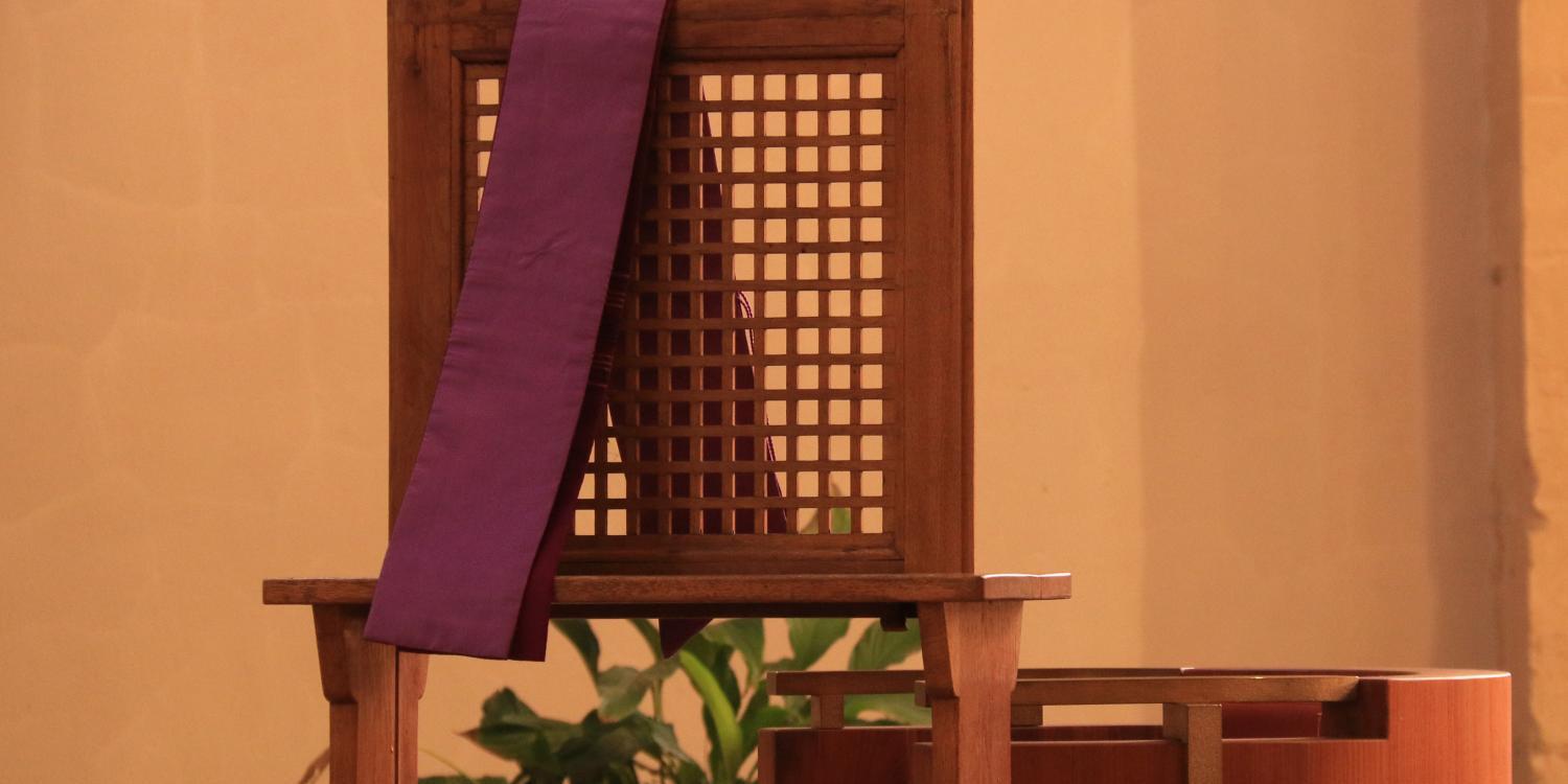 Foto eines Beichtstuhls aus Holz, über der Lehne hängt eine violette Schärpe