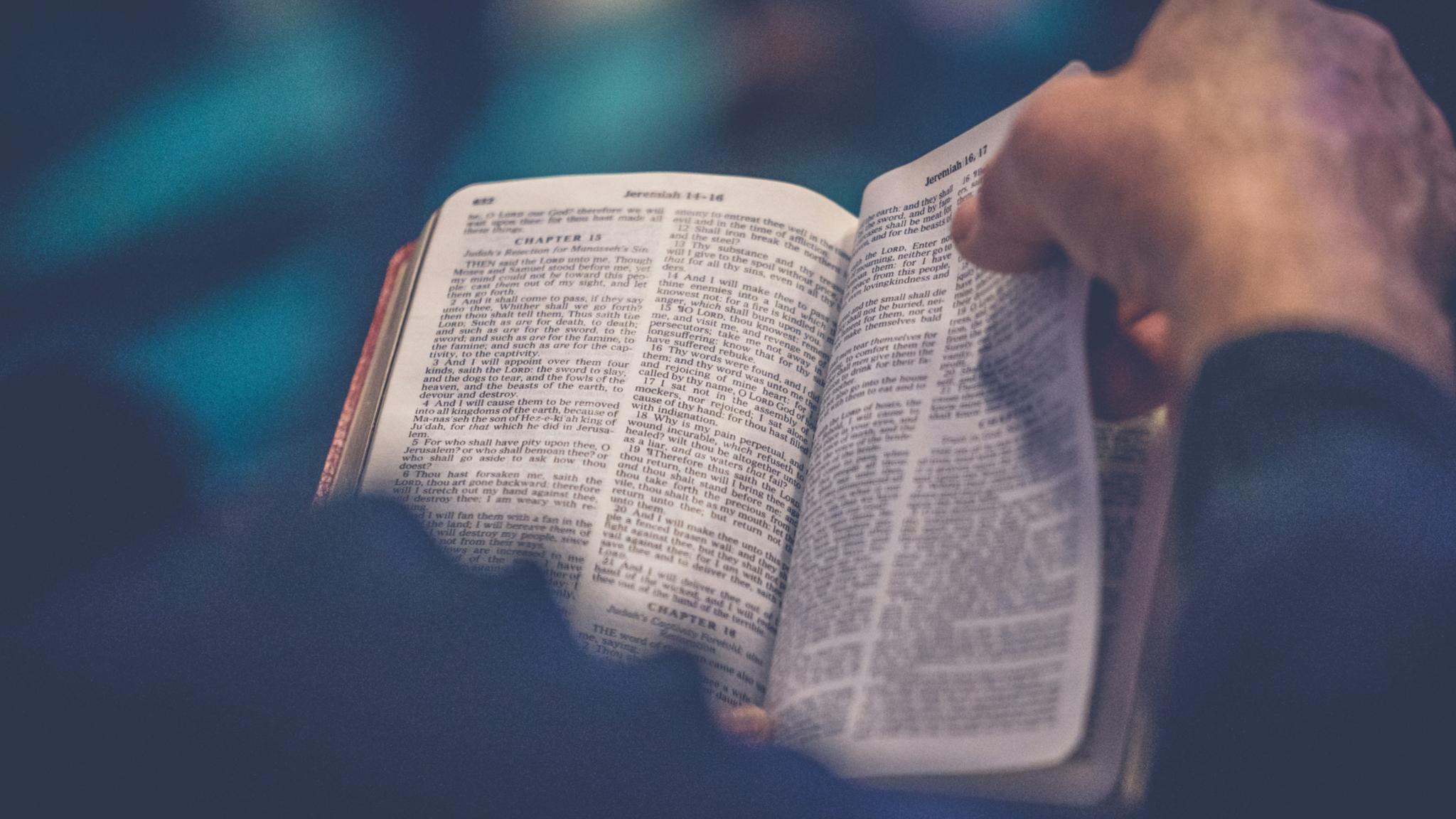 Ein Gebetbuch oder eine Bibel in der eine Person liest, das Buch ist von oben fotografiert, über die rechte Schulter des Lesenden, mit der rechten Hand beginnt die Person gerade, eine Seite umzublättern