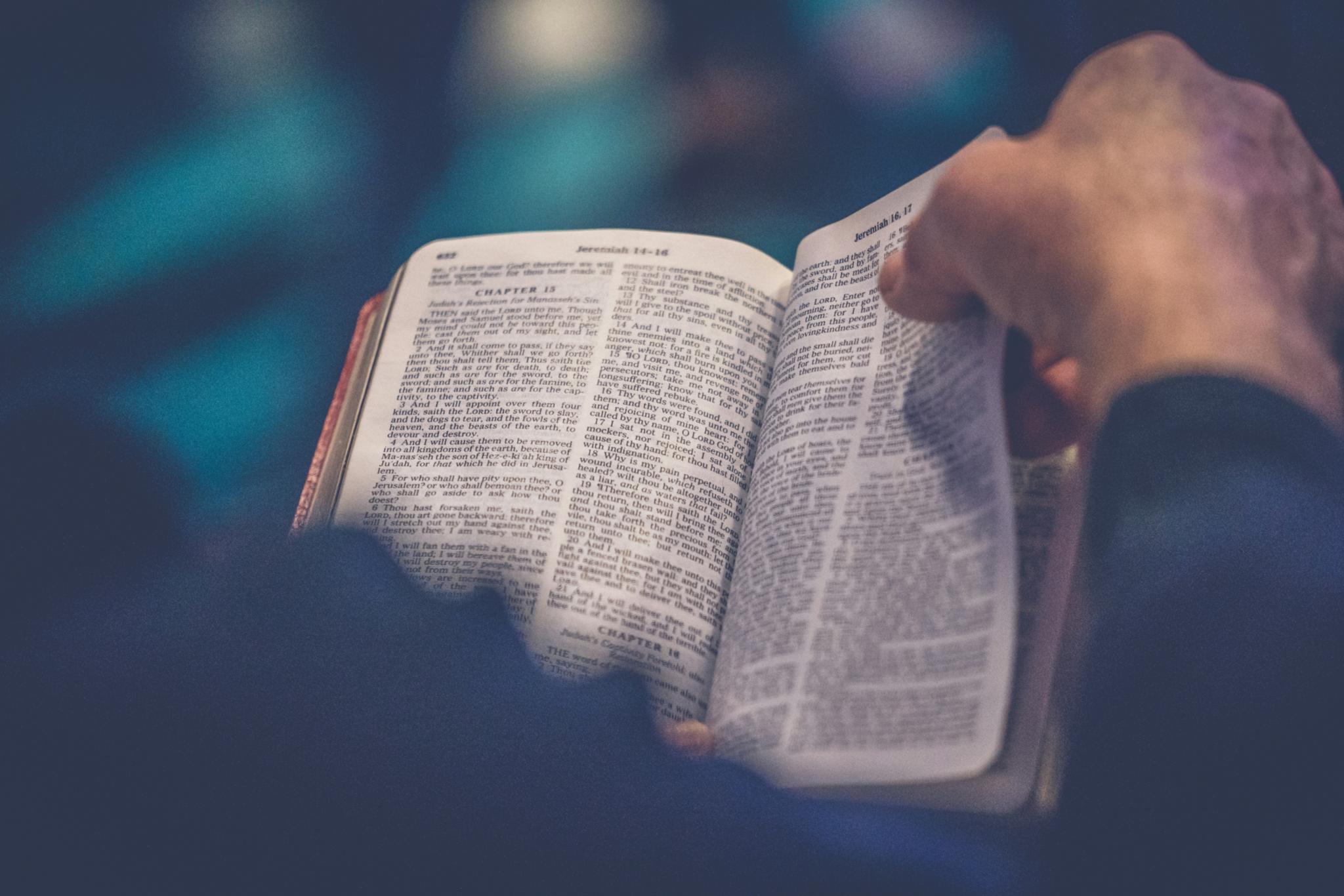 Ein Gebetbuch oder eine Bibel in der eine Person liest, das Buch ist von oben fotografiert, über die rechte Schulter des Lesenden, mit der rechten Hand beginnt die Person gerade, eine Seite umzublättern