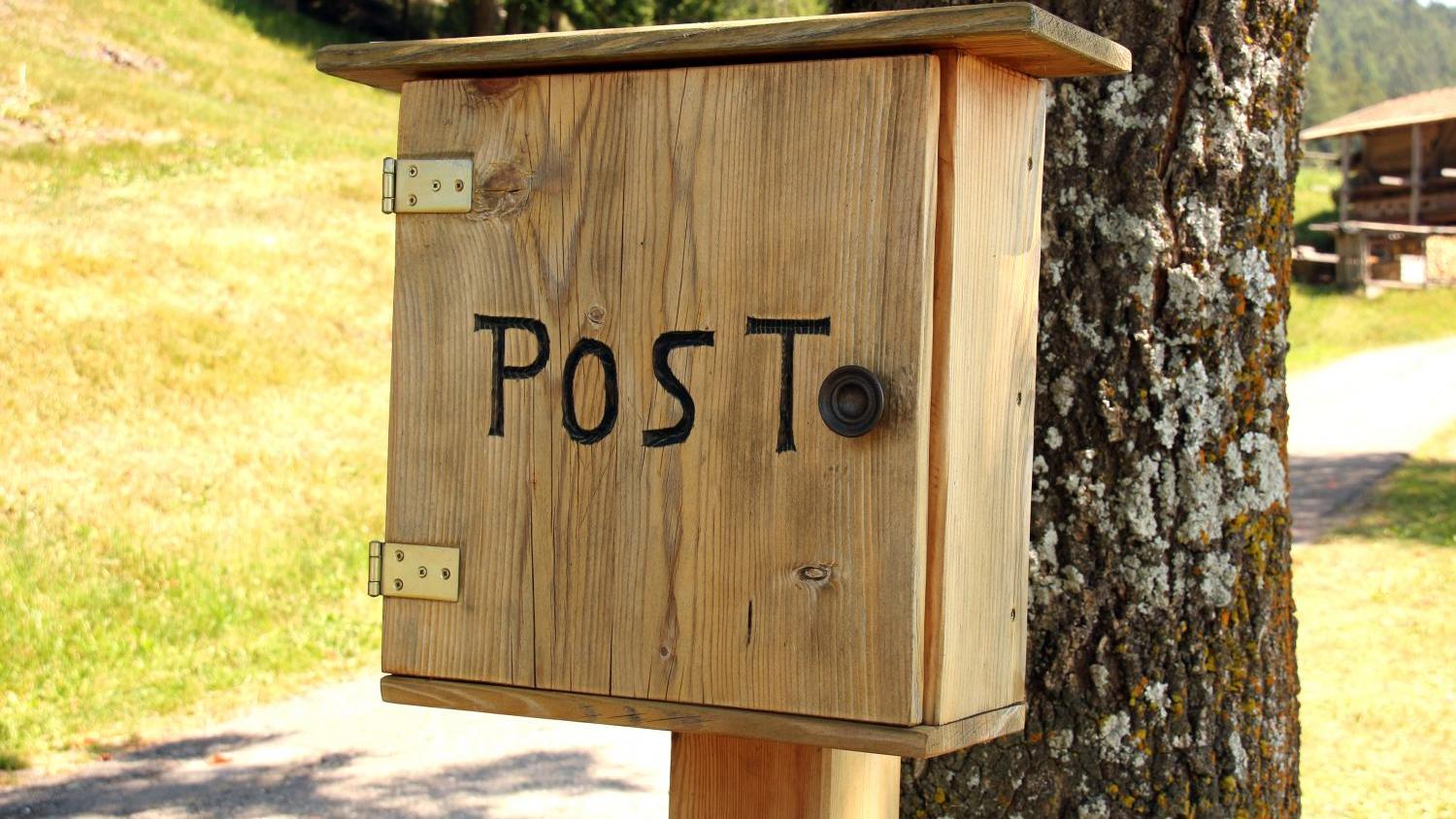 Holzbriefkasten mit der Aufschrift POST der vor einem Baumstamm befestigt ist; dahinter ist ein Feldweg an einem Berghang zu sehen