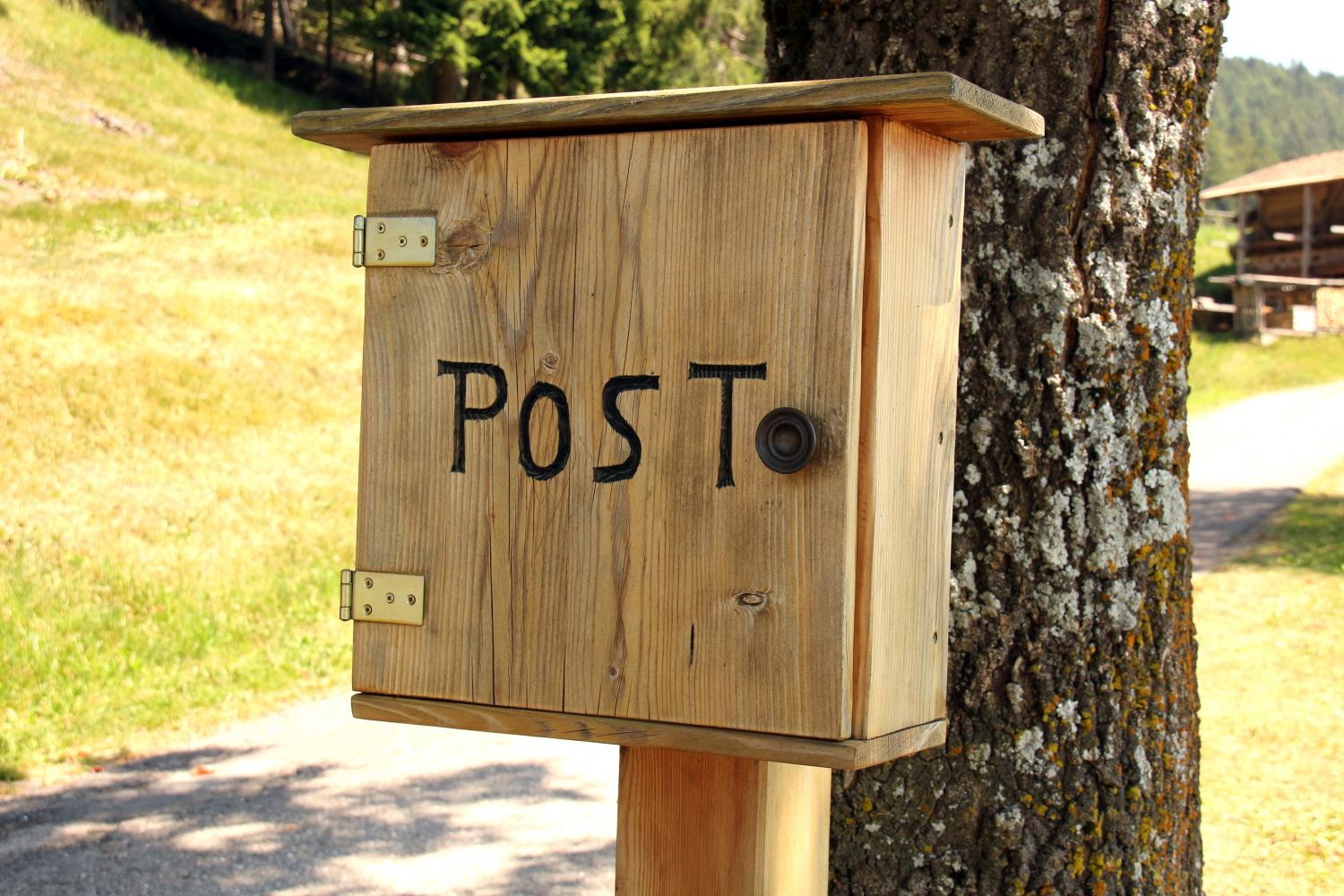 Holzbriefkasten mit der Aufschrift POST der vor einem Baumstamm befestigt ist; dahinter ist ein Feldweg an einem Berghang zu sehen