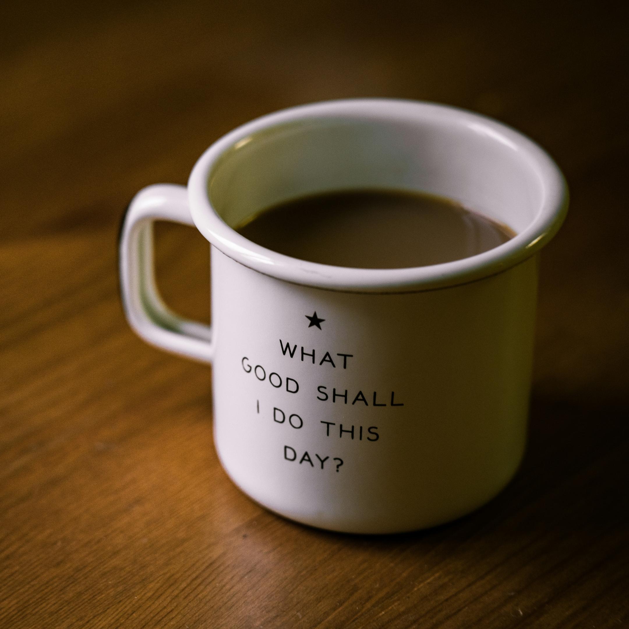 Weisse Tasse auf braunem Tisch; Aufschrift auf der Tasse: 'What Good Shall I Do This Day?'