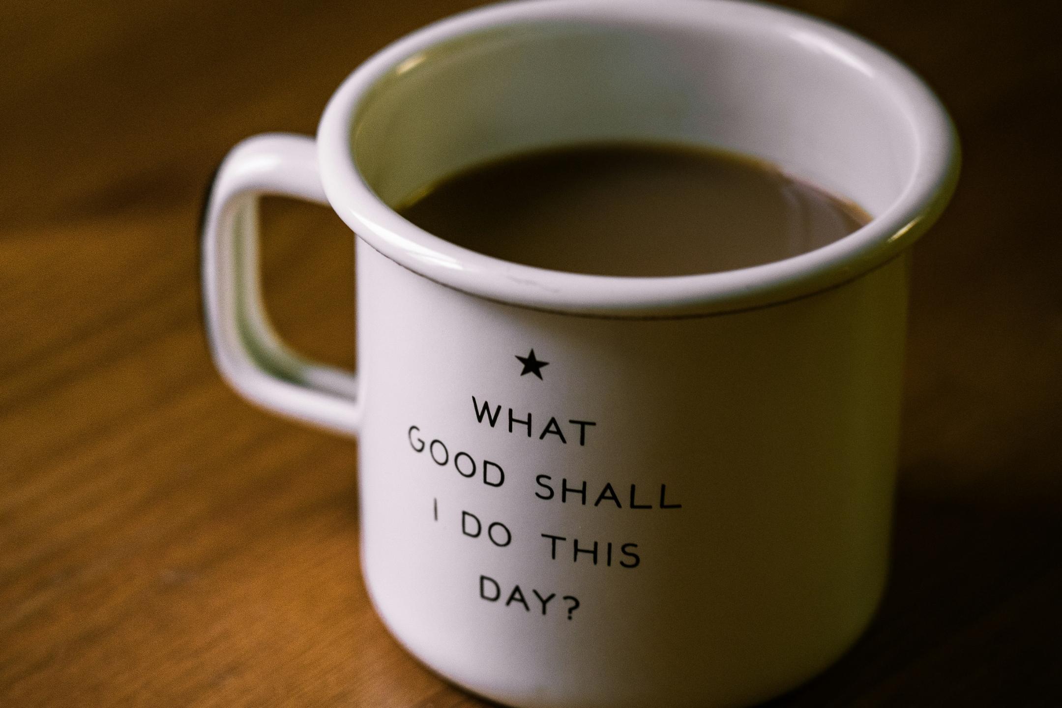 Weisse Tasse auf braunem Tisch; Aufschrift auf der Tasse: 'What Good Shall I Do This Day?'