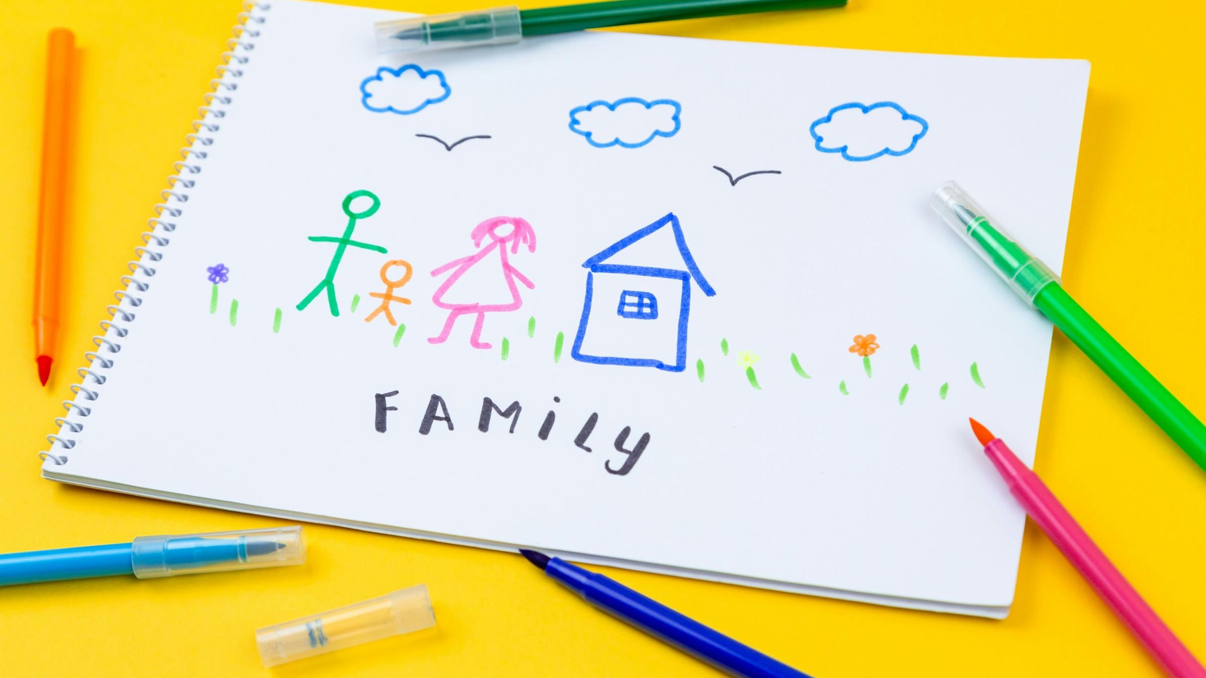 ein Spiralheft liegt auf einem gelben Tisch; drumherum sind Buntstifte verteilt; auf dem obersten Blatt sind - vermutlich von einem Kind - eine Familie als Strichmännchen, ein Haus, Wolken und Blumen gemalt; darunter steht 'Family'