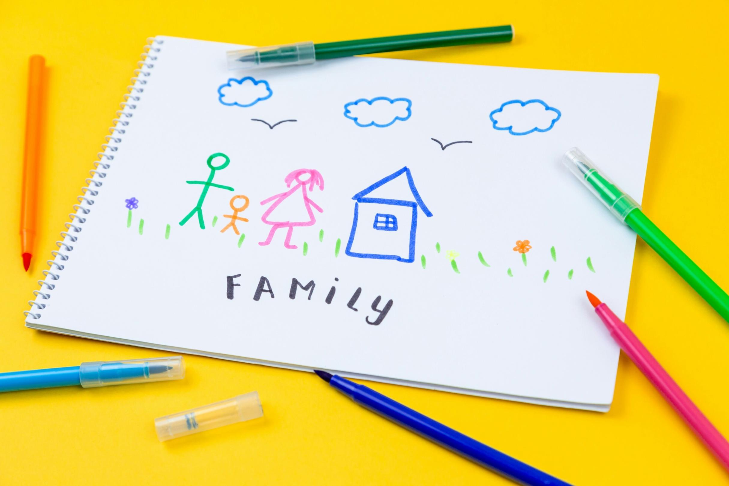 ein Spiralheft liegt auf einem gelben Tisch; drumherum sind Buntstifte verteilt; auf dem obersten Blatt sind - vermutlich von einem Kind - eine Familie als Strichmännchen, ein Haus, Wolken und Blumen gemalt; darunter steht 'Family'