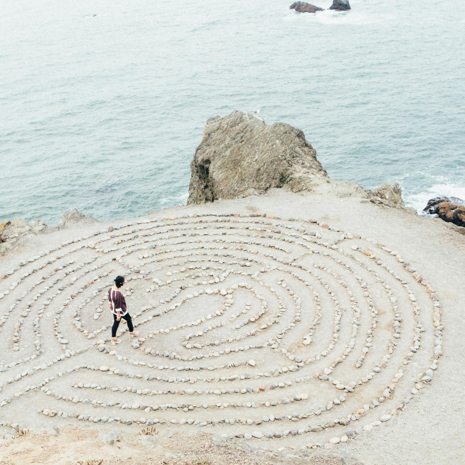 ein kreisrundes Labyrinth aus hellen Steinen auf hellem  Sand direkt an der Küste, zwei kleinere Felsen bilden die Grenze zwischen Wasser und Land, auf oder in dem Labyrinth geht eine junge Frau