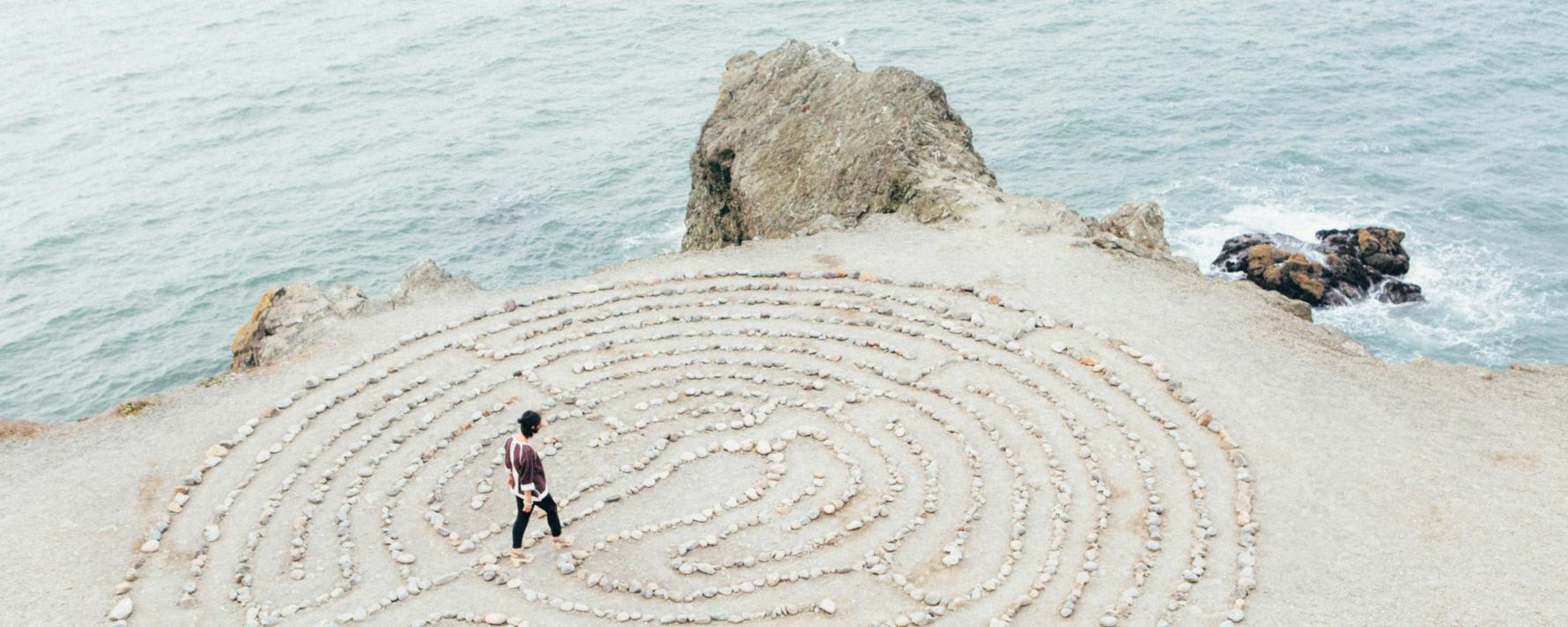 ein kreisrundes Labyrinth aus hellen Steinen auf hellem  Sand direkt an der Küste, zwei kleinere Felsen bilden die Grenze zwischen Wasser und Land, auf oder in dem Labyrinth geht eine junge Frau