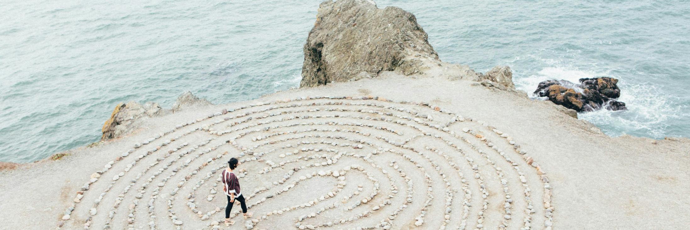 ein kreisrundes Labyrinth aus hellen Steinen auf hellem  Sand direkt an der Küste, zwei kleinere Felsen bilden die Grenze zwischen Wasser und Land, auf oder in dem Labyrinth geht eine junge Frau