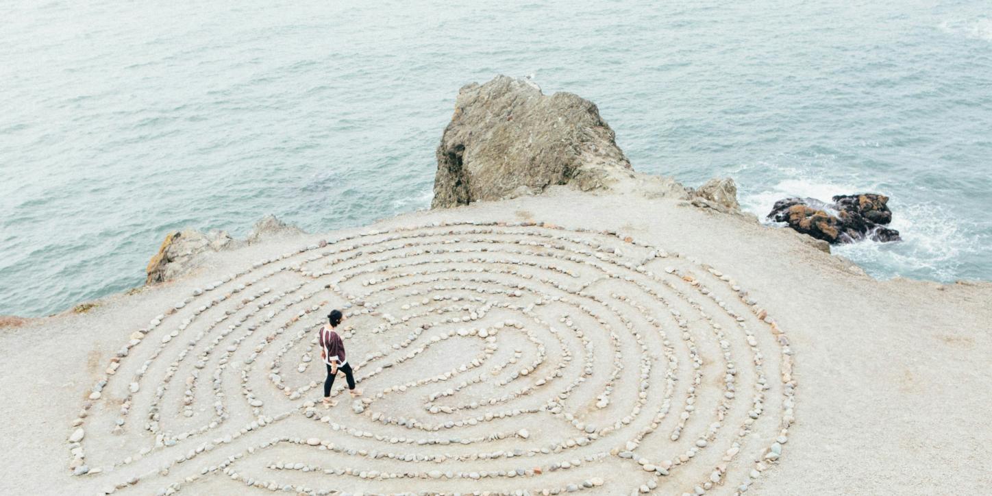 ein kreisrundes Labyrinth aus hellen Steinen auf hellem  Sand direkt an der Küste, zwei kleinere Felsen bilden die Grenze zwischen Wasser und Land, auf oder in dem Labyrinth geht eine junge Frau