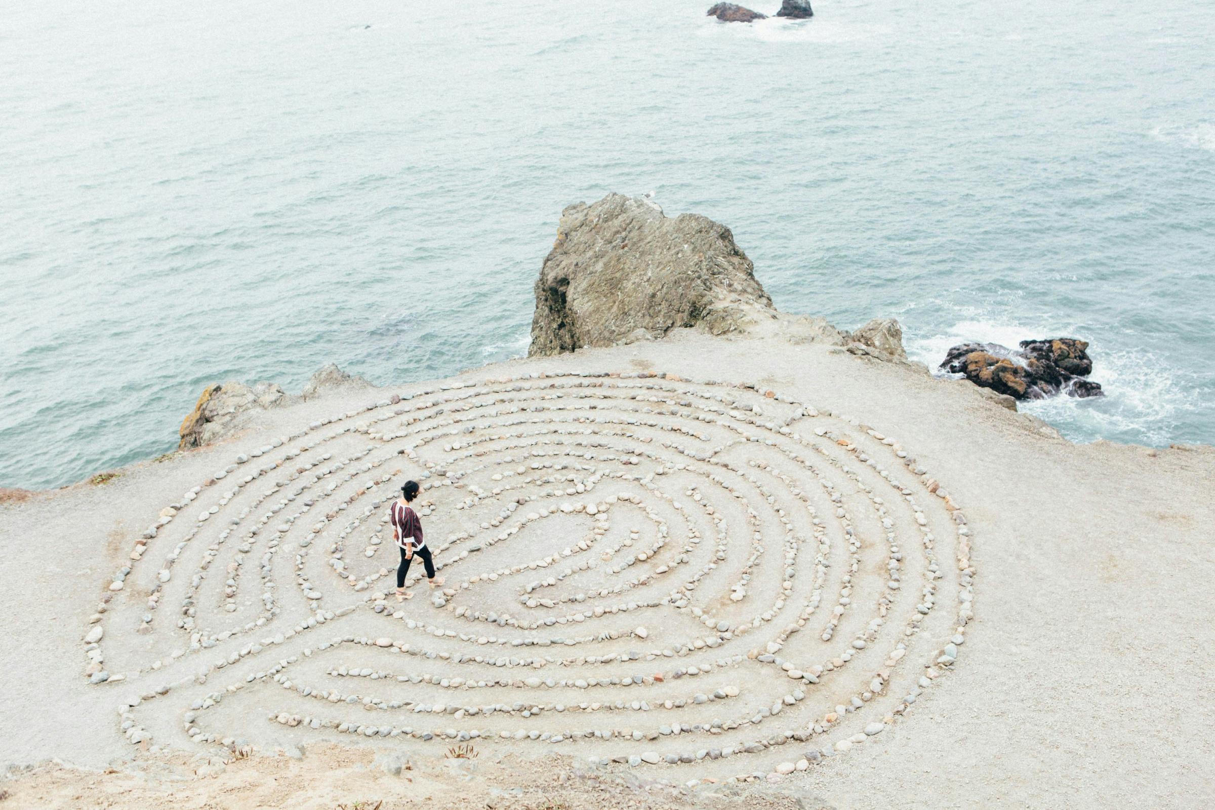 ein kreisrundes Labyrinth aus hellen Steinen auf hellem  Sand direkt an der Küste, zwei kleinere Felsen bilden die Grenze zwischen Wasser und Land, auf oder in dem Labyrinth geht eine junge Frau