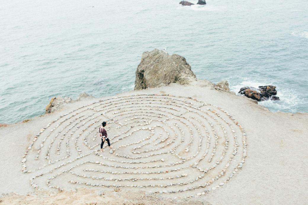 ein kreisrundes Labyrinth aus hellen Steinen auf hellem  Sand direkt an der Küste, zwei kleinere Felsen bilden die Grenze zwischen Wasser und Land, auf oder in dem Labyrinth geht eine junge Frau