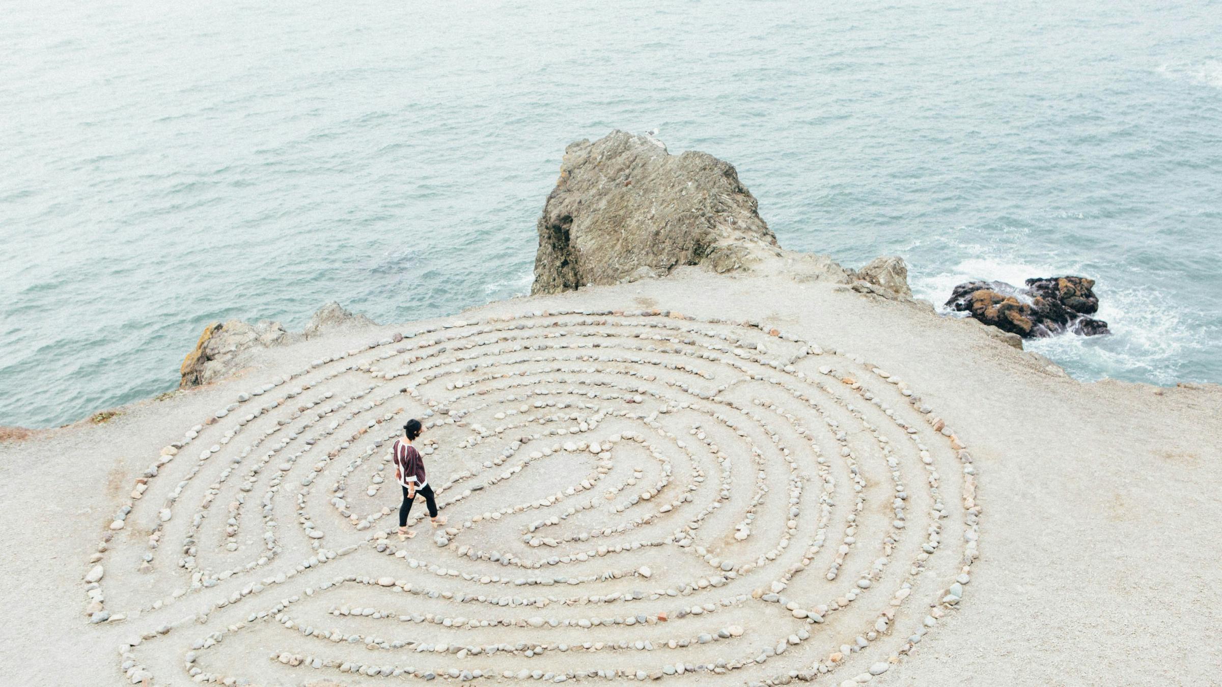 ein kreisrundes Labyrinth aus hellen Steinen auf hellem  Sand direkt an der Küste, zwei kleinere Felsen bilden die Grenze zwischen Wasser und Land, auf oder in dem Labyrinth geht eine junge Frau