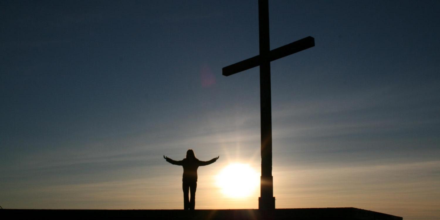 Vor einem Sonnenuntergang als Schatten zu erkennender Mensch neben einem großen Kreuz; der Mensch breitet die Arme aus