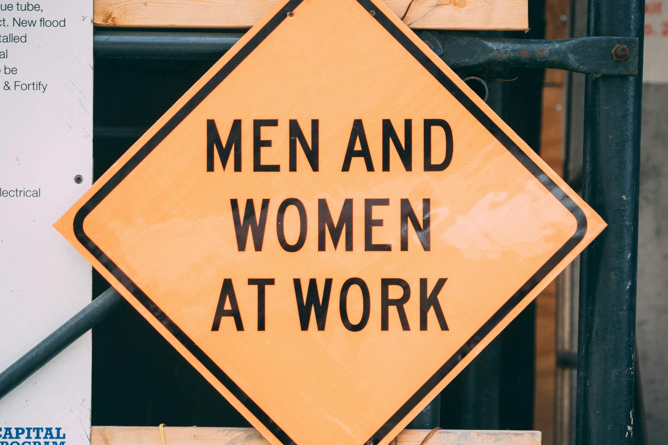 Quadratisches Schild, gelb mit schwarzem Rand, als Raute angeordnet, mit schwarz gehaltener Aufschrift 'Men and Women at Work'