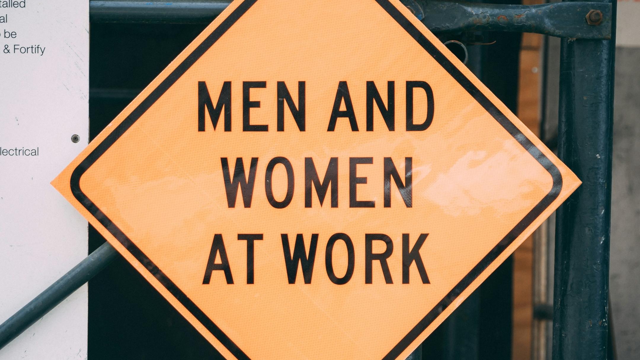 Quadratisches Schild, gelb mit schwarzem Rand, als Raute angeordnet, mit schwarz gehaltener Aufschrift 'Men and Women at Work'