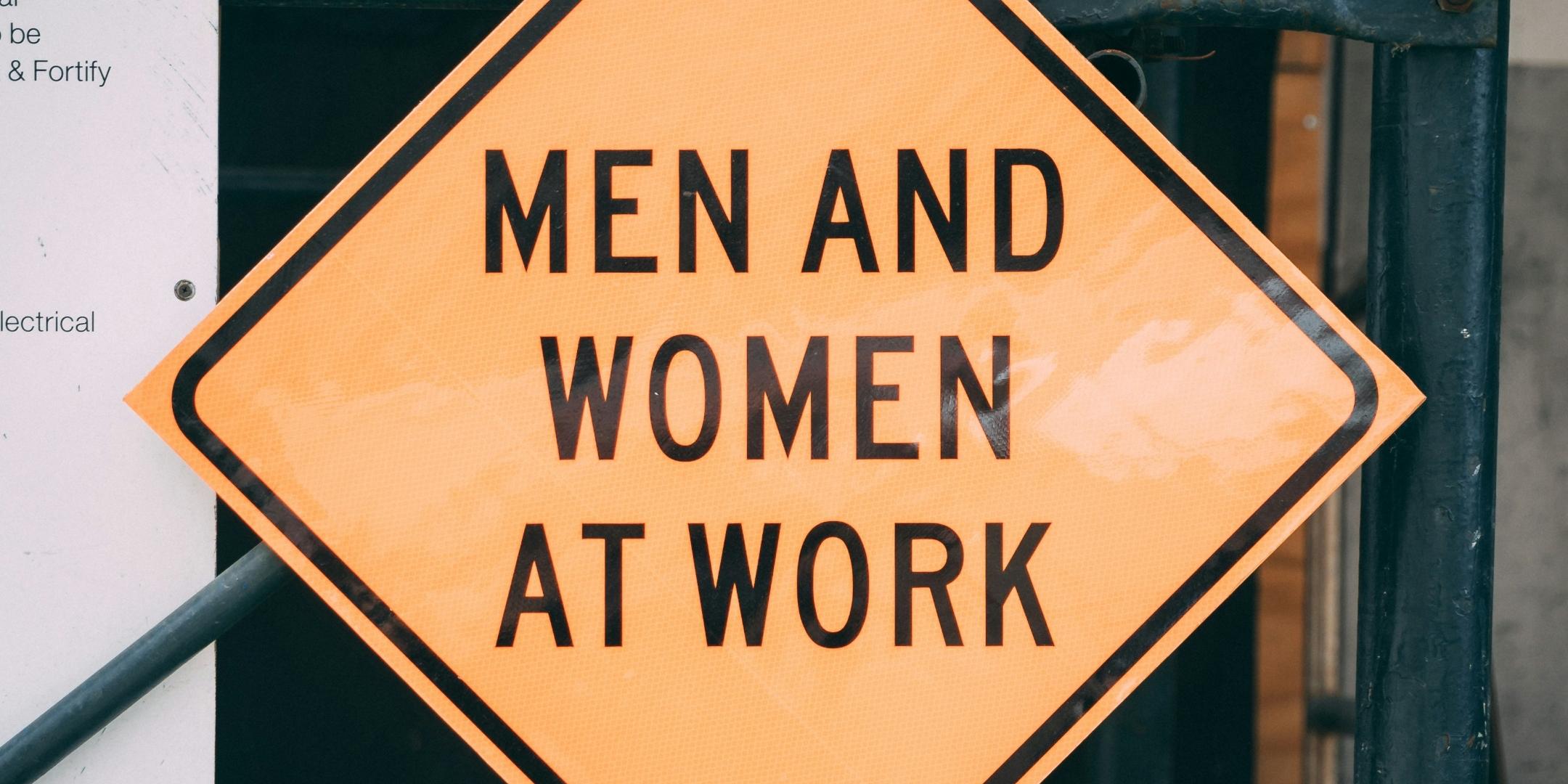Quadratisches Schild, gelb mit schwarzem Rand, als Raute angeordnet, mit schwarz gehaltener Aufschrift 'Men and Women at Work'