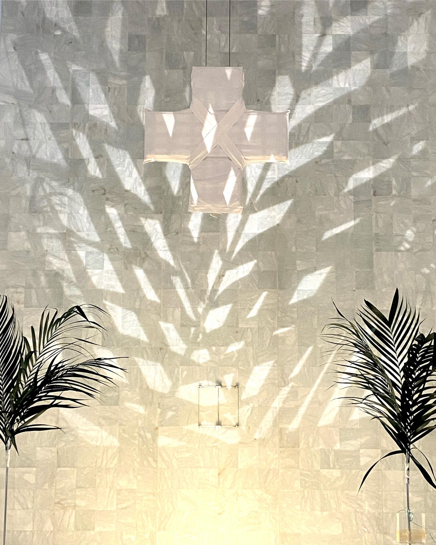 links und rechts die Spitzen von zwei Palmenzweigen die Schatten an eine Wand werfen, vor der ein weißes Kreuz hängt