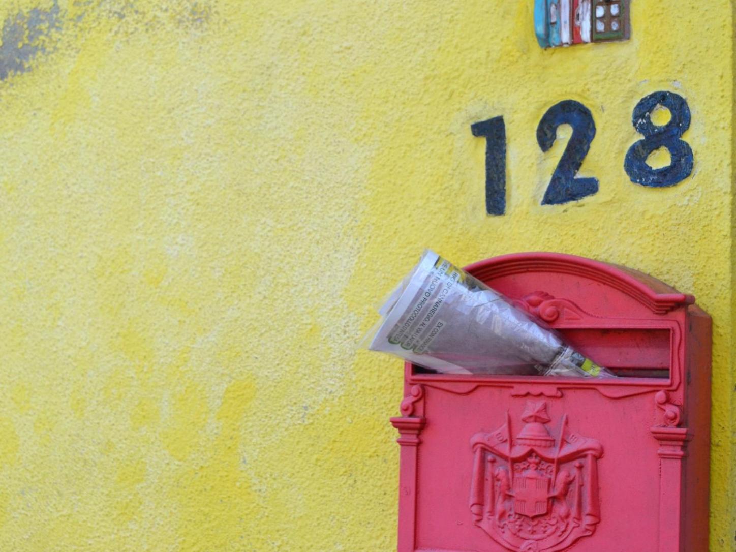 Zeitschrift steckt in einem roten, südeuropäisch anmutenden Briefkasten an gelber Hauswand