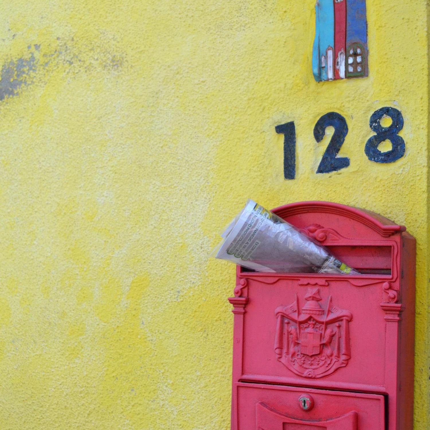 Zeitschrift steckt in einem roten, südeuropäisch anmutenden Briefkasten an gelber Hauswand