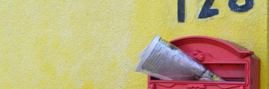 Zeitschrift steckt in einem roten, südeuropäisch anmutenden Briefkasten an gelber Hauswand
