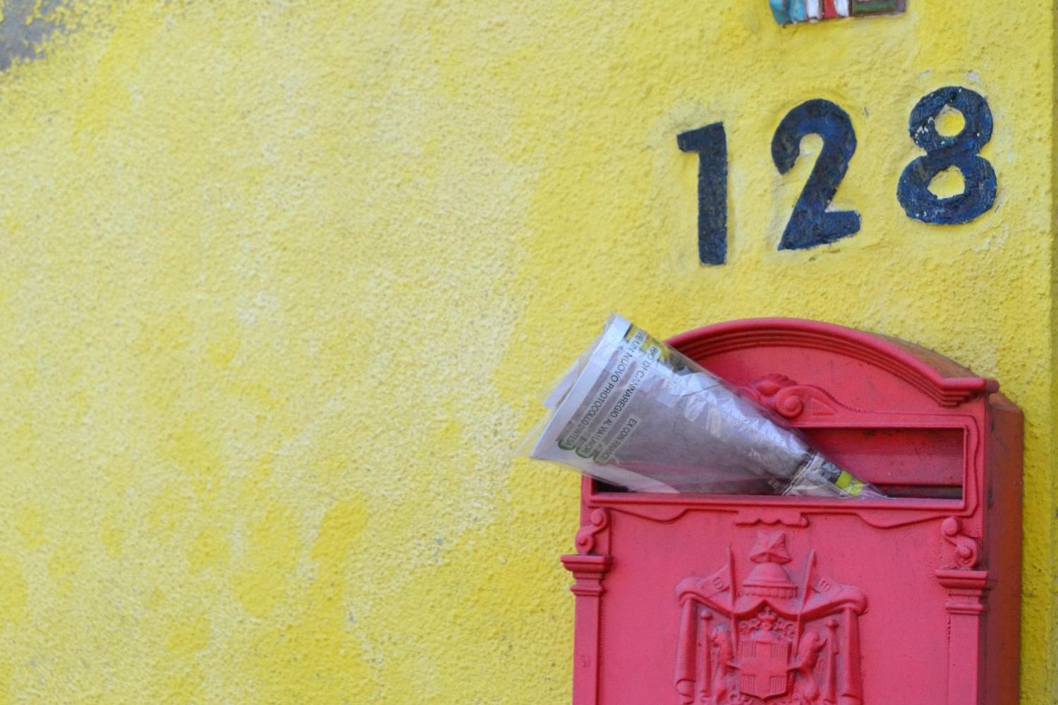 Zeitschrift steckt in einem roten, südeuropäisch anmutenden Briefkasten an gelber Hauswand