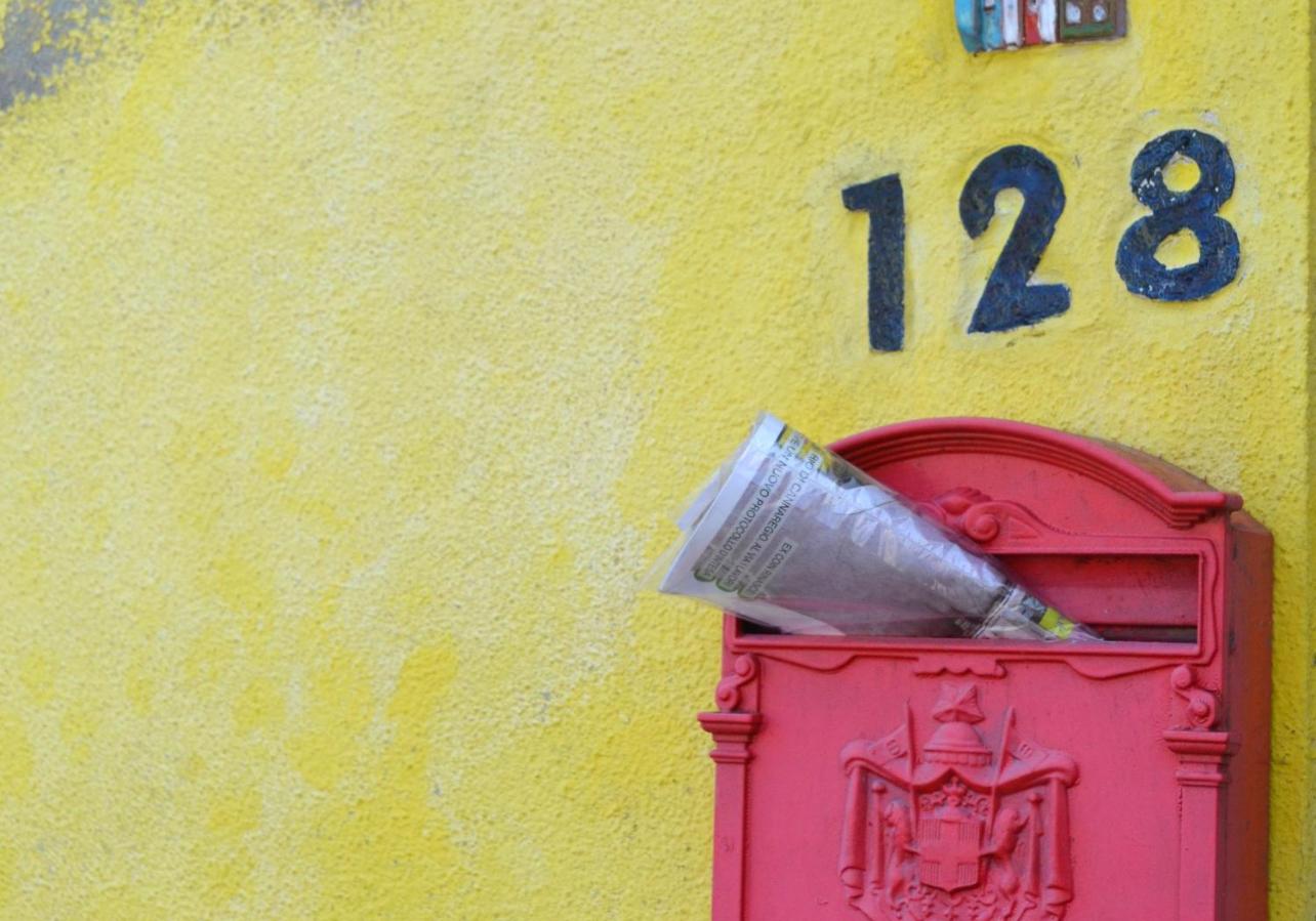 Zeitschrift steckt in einem roten, südeuropäisch anmutenden Briefkasten an gelber Hauswand