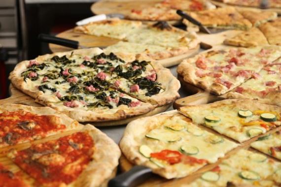 Das Foto zeigt mehrere verschiedene, bereits fertig gebackene Pizzen auf einem Tisch, im Hintergrund sieht man Teile der Ausstattung einer (professionellen) Küche