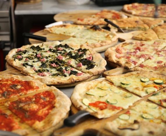 Das Foto zeigt mehrere verschiedene, bereits fertig gebackene Pizzen auf einem Tisch, im Hintergrund sieht man Teile der Ausstattung einer (professionellen) Küche