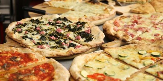 Das Foto zeigt mehrere verschiedene, bereits fertig gebackene Pizzen auf einem Tisch, im Hintergrund sieht man Teile der Ausstattung einer (professionellen) Küche
