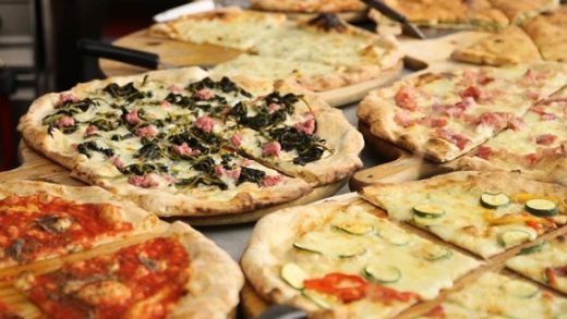 Das Foto zeigt mehrere verschiedene, bereits fertig gebackene Pizzen auf einem Tisch, im Hintergrund sieht man Teile der Ausstattung einer (professionellen) Küche